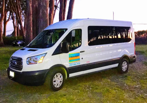 A white passenger van with Mendo Insider Tours branding parked on a grassy area near tall trees.
