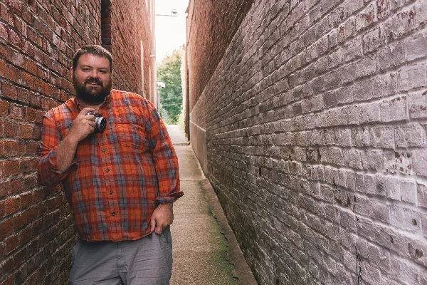 A bearded man in a plaid shirt holding a camera, standing in a brick alleyway.