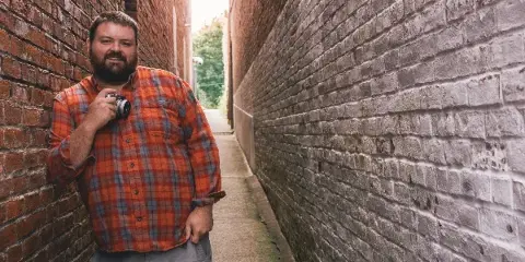 A bearded man in a plaid shirt holding a camera, standing in a brick alleyway.