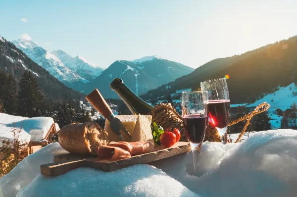 A charcuterie board with cheese, bread, and wine glasses set on a snowy ledge with a mountain valley in the background.
