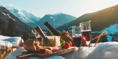 A charcuterie board with cheese, bread, and wine glasses set on a snowy ledge with a mountain valley in the background.