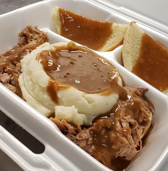A white takeout container holding pulled meat, mashed potatoes, and bread slices, all topped with brown gravy.