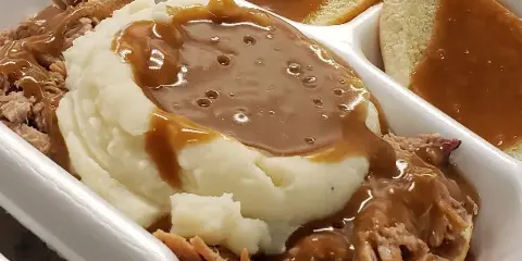 A white takeout container holding pulled meat, mashed potatoes, and bread slices, all topped with brown gravy.