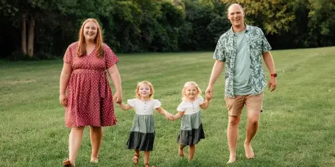 A family of four standing on a grassy field holding hands, captured in a professional outdoor portrait.