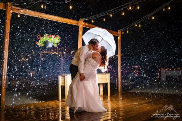 A bride and groom dancing on a wooden deck at night under string lights with rain falling.