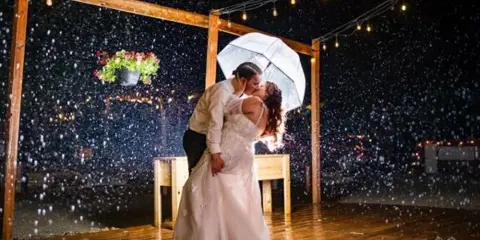 A bride and groom dancing on a wooden deck at night under string lights with rain falling.