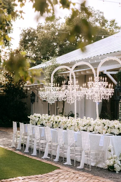 A long outdoor wedding reception table set with white linens, crystal chiavari chairs, and large white floral centerpieces under a white tent structure.