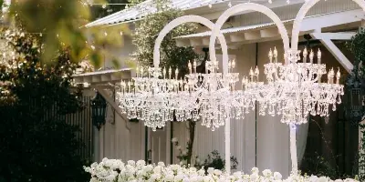 A long outdoor wedding reception table set with white linens, crystal chiavari chairs, and large white floral centerpieces under a white tent structure.