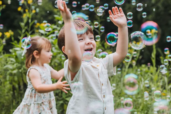 A young boy and girl laughing and reaching for floating soap bubbles in a garden setting.