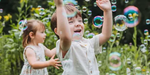 A young boy and girl laughing and reaching for floating soap bubbles in a garden setting.