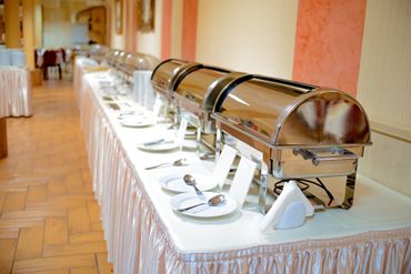 A long catering table set with multiple stainless steel chafing dishes and white plates in a banquet hall.
