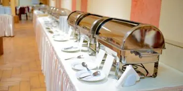 A long catering table set with multiple stainless steel chafing dishes and white plates in a banquet hall.
