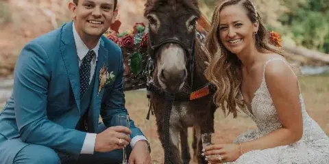 A bride and groom in wedding attire crouching outdoors with a donkey in a mountain landscape.