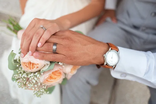 Close-up of a bride and groom's hands holding a bouquet of peach roses and baby's breath, showing wedding rings.
