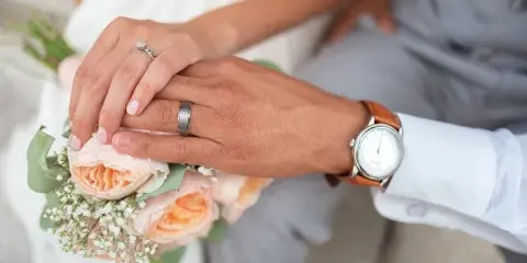 Close-up of a bride and groom's hands holding a bouquet of peach roses and baby's breath, showing wedding rings.