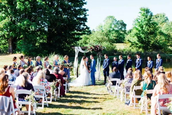 An outdoor wedding ceremony with a bride and groom at an altar, surrounded by a wedding party and seated guests in a grassy field.