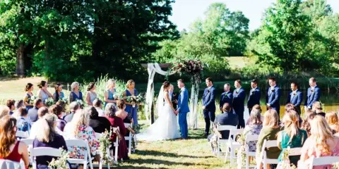 An outdoor wedding ceremony with a bride and groom at an altar, surrounded by a wedding party and seated guests in a grassy field.
