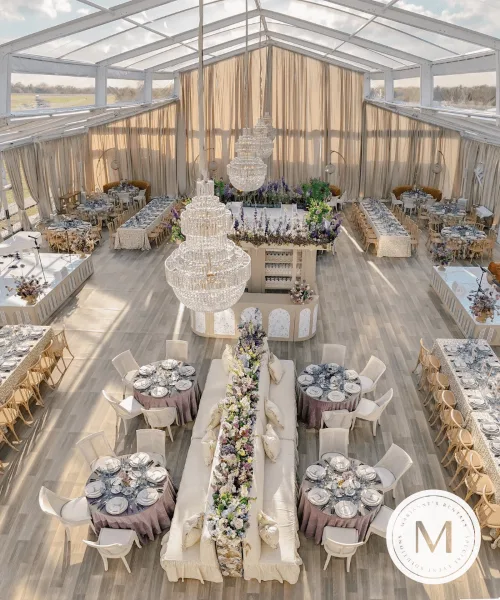 Interior view of a clear-top event tent featuring chandeliers, lounge seating, and multiple dining tables set with tableware.