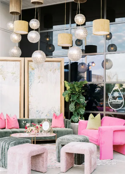 A styled event lounge with green and pink velvet sofas, poufs, a coffee table, and hanging pendant lights.