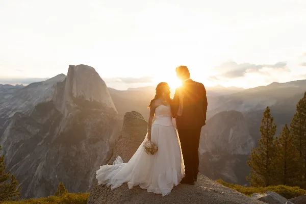 A bride and groom standing on a rocky outcrop overlooking a mountain valley at sunset.