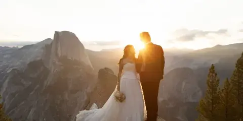 A bride and groom standing on a rocky outcrop overlooking a mountain valley at sunset.