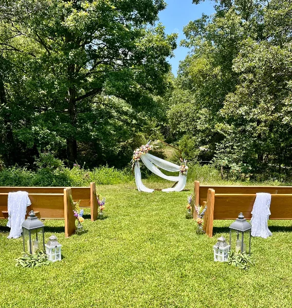 Outdoor wedding ceremony setup with a floral arch, wooden benches, and lanterns on a grassy lawn.