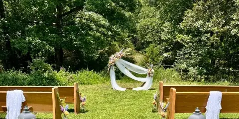 Outdoor wedding ceremony setup with a floral arch, wooden benches, and lanterns on a grassy lawn.