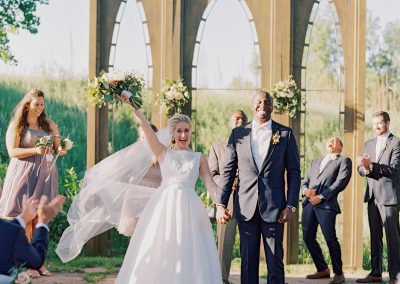 A bride and groom with their wedding party celebrating in front of an arched outdoor pavilion structure.