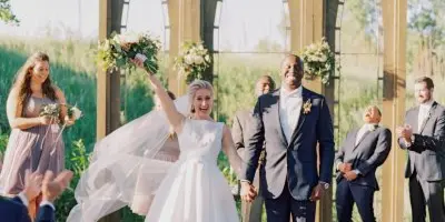 A bride and groom with their wedding party celebrating in front of an arched outdoor pavilion structure.