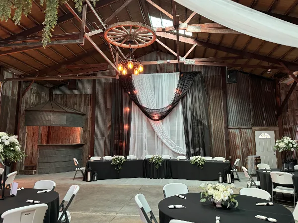 Interior of a rustic wedding venue featuring a high wooden ceiling, a hanging wagon wheel, and a decorated head table with white and black drapery.