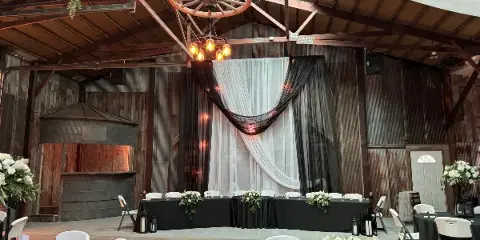Interior of a rustic wedding venue featuring a high wooden ceiling, a hanging wagon wheel, and a decorated head table with white and black drapery.