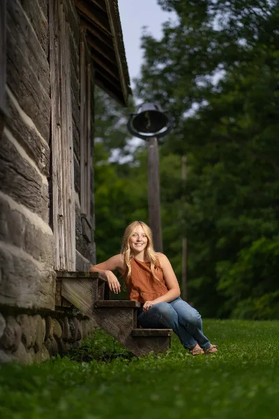 A young woman with long blonde hair in an orange top and jeans sits on a wooden step beside a log cabin in a grassy, wooded area.