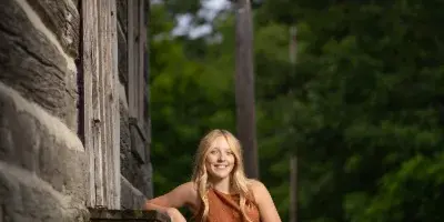 A young woman with long blonde hair in an orange top and jeans sits on a wooden step beside a log cabin in a grassy, wooded area.