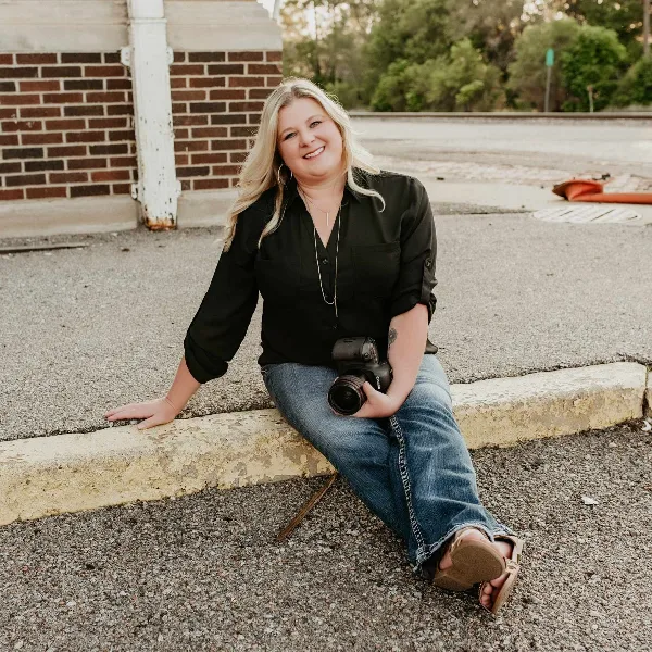 A smiling woman with blonde hair sitting on a curb holding a professional camera.