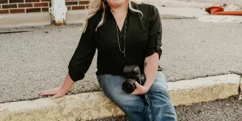 A smiling woman with blonde hair sitting on a curb holding a professional camera.