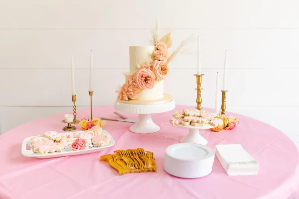 A white two-tier wedding cake decorated with pink flowers and wheat, displayed on a white pedestal stand on a pink tablecloth alongside decorated cookies and gold forks.
