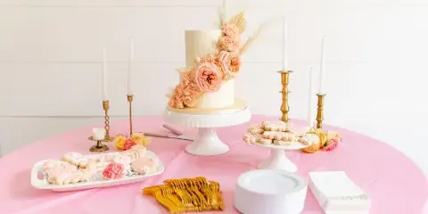 A white two-tier wedding cake decorated with pink flowers and wheat, displayed on a white pedestal stand on a pink tablecloth alongside decorated cookies and gold forks.