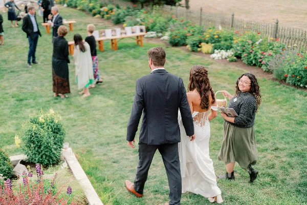 A bride and groom walking through a garden with a wedding planner holding a clipboard and basket.