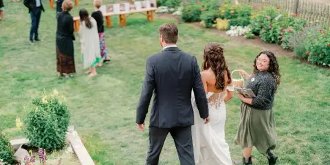 A bride and groom walking through a garden with a wedding planner holding a clipboard and basket.
