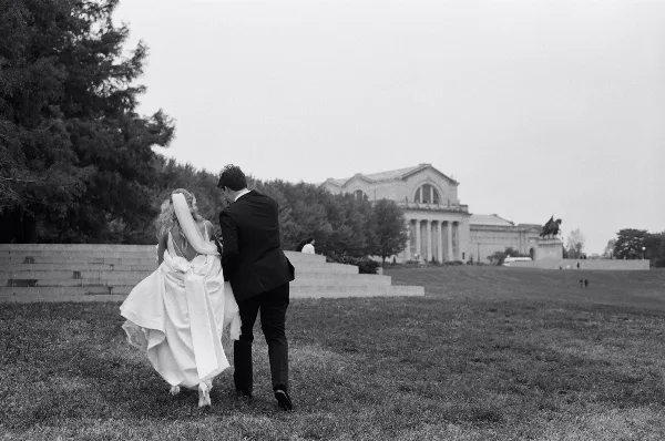 Black and white documentary-style photograph of a bride and groom walking on a grassy lawn with a large classical building in the background.