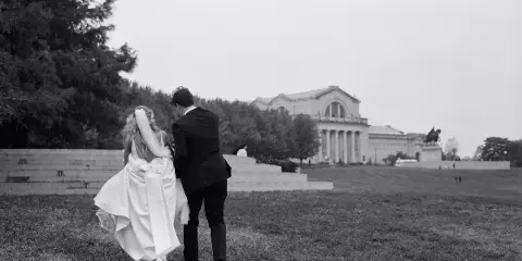 Black and white documentary-style photograph of a bride and groom walking on a grassy lawn with a large classical building in the background.