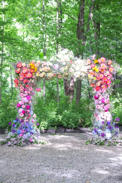 A lush, colorful floral wedding arch featuring pink, yellow, and purple flowers set against a backdrop of green trees.
