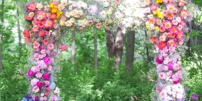 A lush, colorful floral wedding arch featuring pink, yellow, and purple flowers set against a backdrop of green trees.