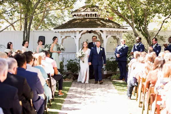 A bride and groom walking down a stone aisle in a garden setting during a wedding ceremony.