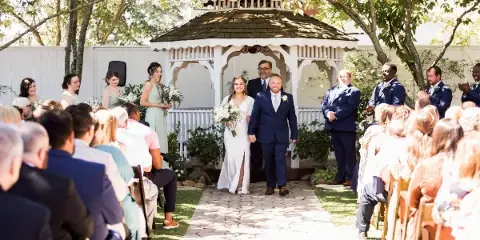 A bride and groom walking down a stone aisle in a garden setting during a wedding ceremony.
