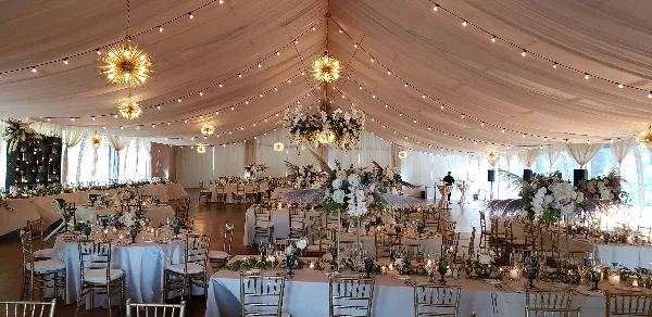 Decorated wedding reception inside a white tent with round tables, chairs, floral centerpieces, and hanging lights.