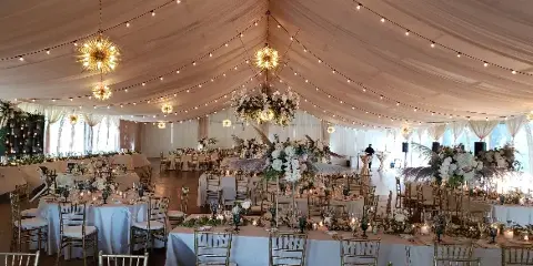 Decorated wedding reception inside a white tent with round tables, chairs, floral centerpieces, and hanging lights.