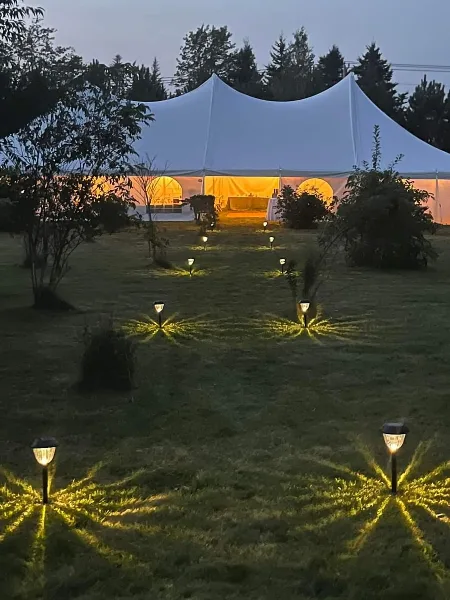 A large white event tent illuminated from within at dusk, with a line of decorative pathway lights leading across a grassy lawn.
