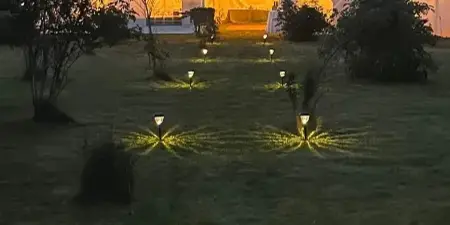 A large white event tent illuminated from within at dusk, with a line of decorative pathway lights leading across a grassy lawn.