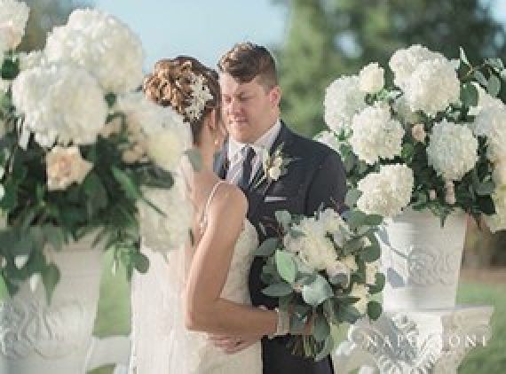 A bride and groom at a wedding surrounded by white floral arrangements and holding a bouquet.